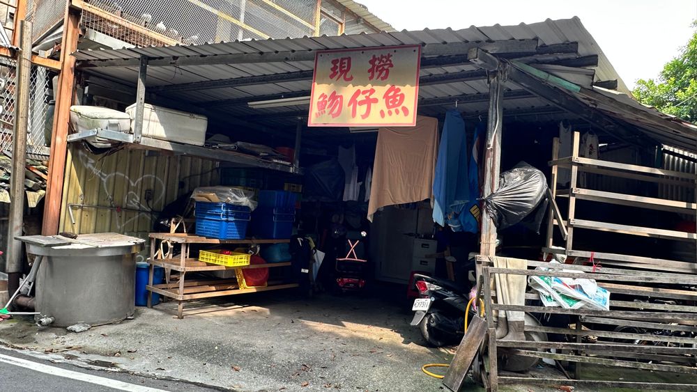 「現撈魩仔魚」招牌高掛在鐵皮棚下，漁具、籃筐與工作檯堆疊其間，是討海人家日常補給的作業空間，也是八里漁村最真實的生活切片。（照片提供者：郭庭禎 / 拍攝日期：2026.08.11）