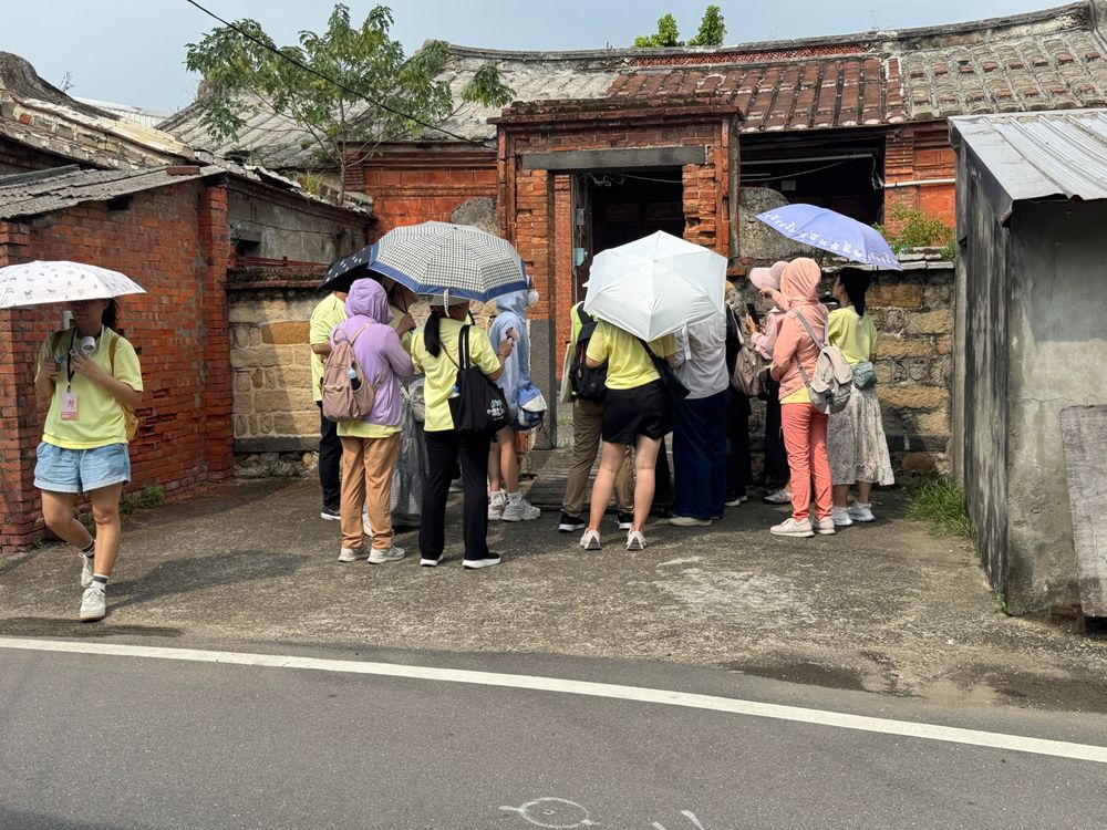 學員圍聚在紅磚古厝門前，聆聽這座百年建築的故事。斑駁的燕尾磚牆與瓦頂，訴說著八里聚落曾經的生活痕跡。（照片提供者：新北市教育局 / 拍攝日期：2026.08.11）